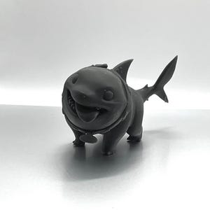 Peut inclure: Une figurine d'un requin en 3D imprim&eacute;e en noir avec un collier de chien et une &eacute;tiquette. Le requin a une expression amicale et est debout sur ses quatre pattes.