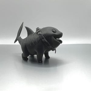 Peut inclure: Une figurine d'un requin en 3D imprim&eacute;e en noir portant un collier. Le requin a un sourire narquois et de grands yeux.