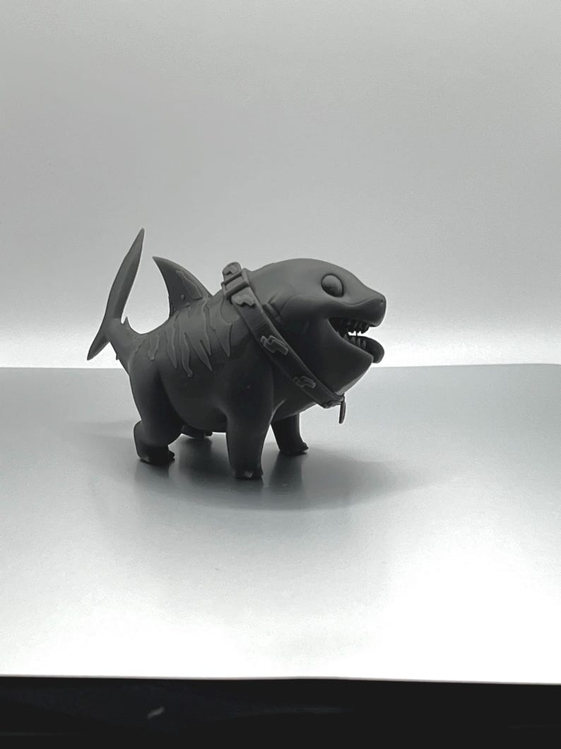Peut inclure: Une figurine d'un requin en 3D imprim&eacute;e en noir portant un collier. Le requin a un sourire narquois et de grands yeux.
