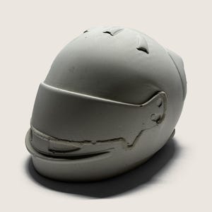 May include: A gray, sculpted helmet with a visor. The helmet has a smooth, matte finish and is designed to resemble a racing helmet. The visor is slightly tinted, and the helmet has details like ear cutouts.