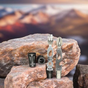 May include: Two silver multi-tool utensils with black handles and two black caps with the text "Trek N' Eat" on them. The tools are on top of a pile of brown rocks.