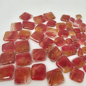 May include: A collection of square, translucent glass tiles in shades of red, orange, and amber. The tiles vary in size and have a marbled appearance, with swirls of color throughout. The edges are rounded, and the tiles are scattered across a white surface.