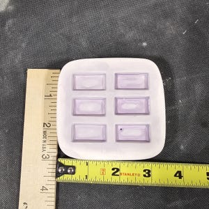 May include: A white silicone mold with six rectangular cavities, positioned next to a wooden ruler and a yellow Stanley measuring tape. The mold measures approximately 2.5 inches in width and 2.5 inches in height, featuring smooth, rounded corners.
