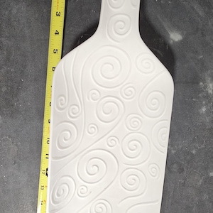 May include: A white ceramic serving platter shaped like a bottle, featuring an embossed swirl pattern across its surface. A yellow measuring tape placed alongside indicates the platter has a total length of approximately 15.5 inches.