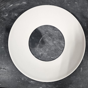 May include: A circular white plate featuring a wide, smooth rim and a recessed, dark gray circular center. The design creates a high-contrast appearance with a clean, matte finish on the outer edge and a textured, darker surface in the middle.