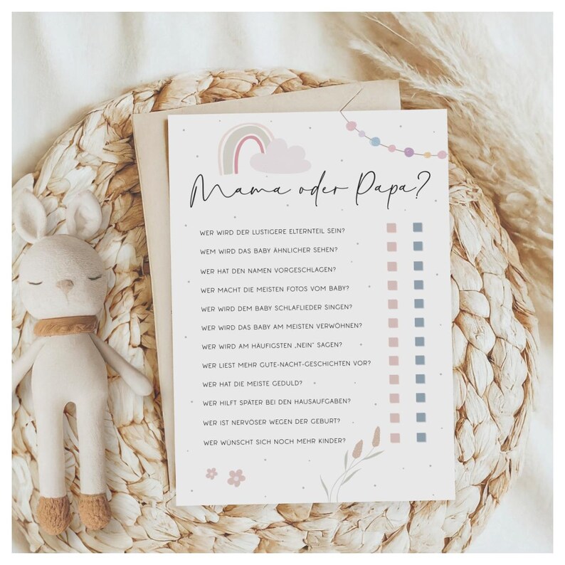 May include: A baby shower game card with the text "Mama oder Papa?" in black script. The card features a rainbow, cloud, and floral design in pastel colours. A stuffed bunny toy with a brown scarf is on the left.