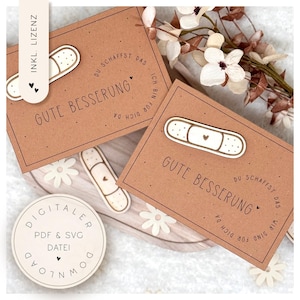 May include: Two brown cards with the words "Gute Besserung" and a bandage design. The cards have a circular text that says "Du schaffst das, ich bin für dich da, wir sind für dich da." A wooden bandage and a round sticker are also visible.
