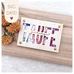 Puede incluir: Un tarjetero de madera de color claro con la palabra "TAUFE" recortada, que revela moneda colorida. La frase "zur heiligen" también está recortada. El soporte está asegurado con remaches dorados y se encuentra sobre una superficie de madera. El texto "SVG DATEI" está en la esquina superior izquierda.