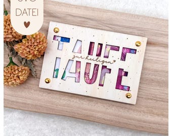 May include: A light-coloured wooden gift card holder with the word "TAUFE" cut out, revealing colourful currency. The phrase "zur heiligen" is also cut out. The holder is secured with gold-coloured rivets and sits on a wooden surface. The text "SVG DATEI" is in the upper left corner.