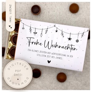 Pode incluir: Uma caixa branca com a inscrição alemã "Frohe Weihnachten" e ornamentos decorativos suspensos. A caixa contém bombons. Um adesivo redondo com texto está no canto inferior esquerdo.