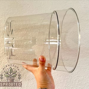 May include: A clear, cylindrical plastic plant pot featuring a wide, open top rim and a series of vertical drainage slits integrated into the base. The transparent material allows for full visibility of the interior and the drainage structure.