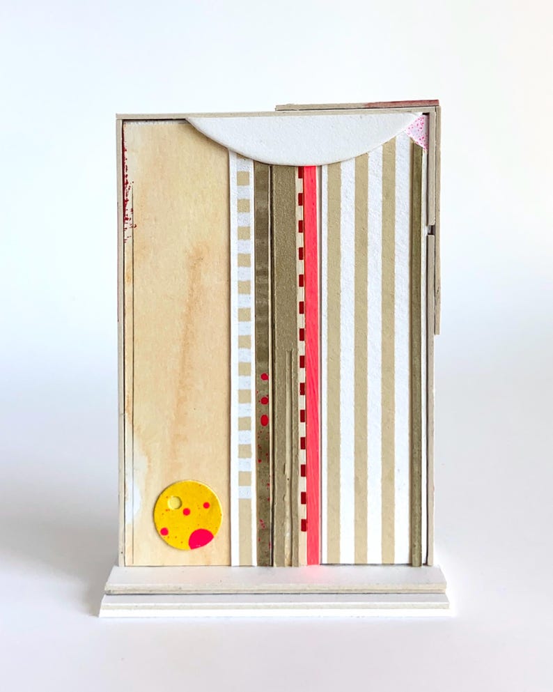 Op de afbeelding: Een mixed media kunstwerk op een rechthoekige houten structuur. Het ontwerp bevat papieren stroken met verticale strepen, een rode lijn en een gele cirkel met roze accenten. Gemonteerd op een witte basis.