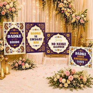 May include: Four decorative signs with text in a wedding setting. The signs are purple and gold with phrases like "Proud Dadke of the Bride" and "Sorry No Nanke Allowed". Floral arrangements and candles surround the signs.