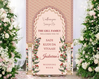 May include: A wedding welcome sign with a light pink background and ornate design. The sign features text in a decorative font, including names and the phrase "Wedding Week." Floral arrangements frame the sign, with white and pink flowers.