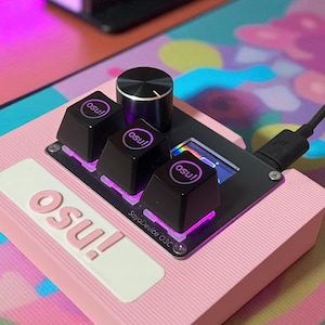 May include: A pink gaming peripheral with the word "osu!" in a raised design. It features black keys with "osu!" printed on them, a silver knob, and a small screen. The device is connected via a USB cable.
