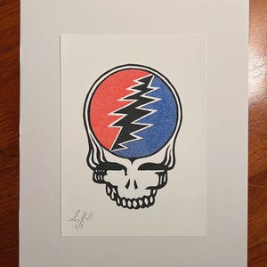 May include: A black and white illustration of a smiling face with a red and blue lightning bolt design inside the head. The artwork is signed "S.K.R.II 5/9".