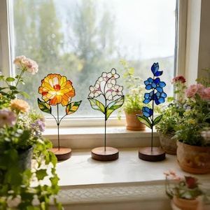 Könnte beinhalten: Drei Glasmalerei-Blumendekorationen. Die erste ist eine gelb-orangefarbene Blume mit grünen Blättern. Die zweite ist eine weiße Blume mit rosa Akzenten und grünen Blättern. Die dritte ist eine blaue Blume mit grünen Blättern. Jede ist auf einem Holzsockel montiert.