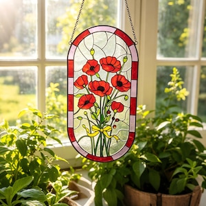 May include: An oval stained glass suncatcher featuring a bouquet of vibrant red poppies with green stems and leaves, tied with a yellow ribbon. The piece is framed by a border of alternating red and light pink glass segments and hangs by a metal chain.