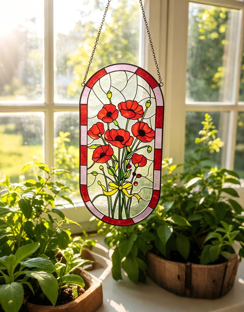 May include: An oval stained glass suncatcher featuring a bouquet of vibrant red poppies with green stems and leaves, tied with a yellow ribbon. The piece is framed by a border of alternating red and light pink glass segments and hangs by a metal chain.