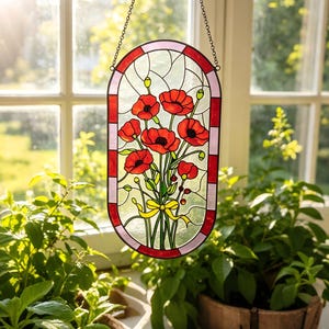 May include: An oval stained glass suncatcher featuring a bouquet of vibrant red poppies with green stems and leaves, tied with a yellow ribbon. The piece is framed by a border of alternating red and light pink glass segments and hangs by a metal chain.