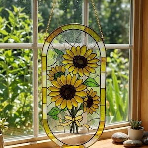 May include: An oval stained glass suncatcher featuring three yellow sunflowers with dark brown centers, green leaves, and a yellow ribbon tied at the stems. The piece is framed in a yellow border and hangs from a gold-colored metal chain.