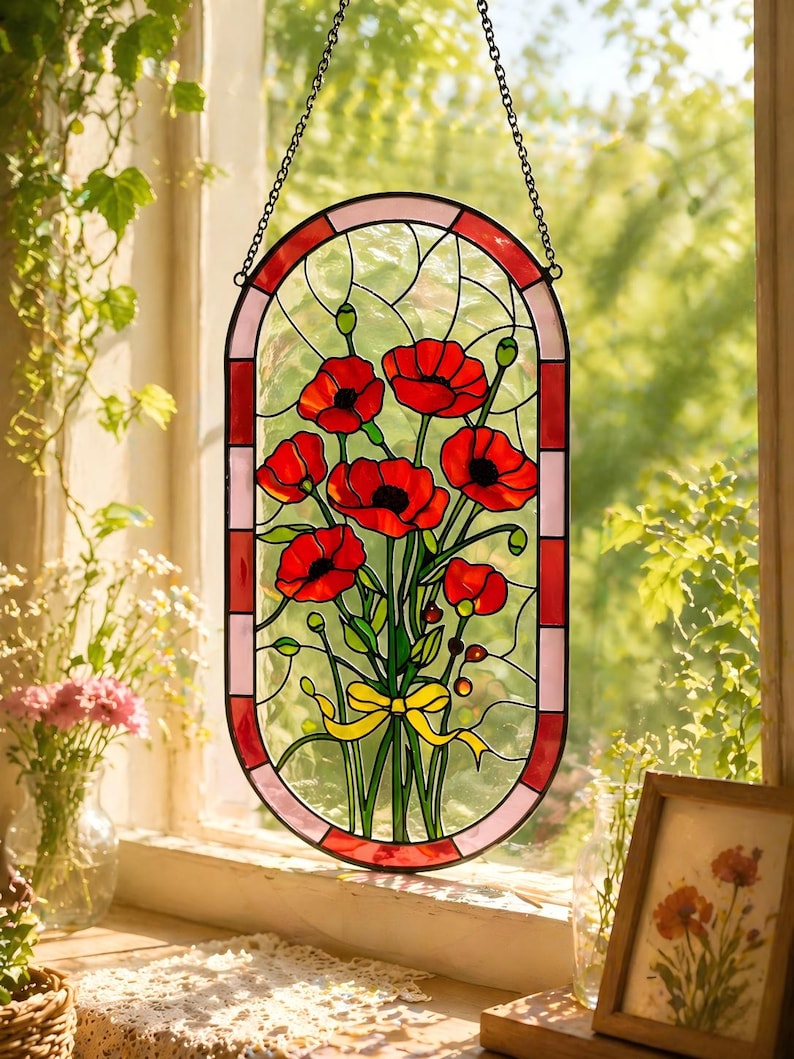 May include: An oval stained glass suncatcher featuring a bouquet of vibrant red poppies with green stems and leaves, tied with a yellow ribbon. The piece is framed by a border of alternating red and light pink glass segments and hangs by a metal chain.