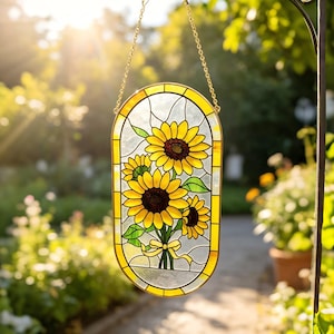 May include: An oval stained glass suncatcher featuring three yellow sunflowers with dark brown centers and green leaves, tied with a yellow ribbon. The piece is framed in a gold-toned metal border and hangs from a gold chain on a metal hook.