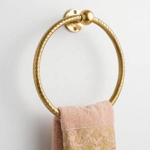 May include: A gold-colored towel ring with a textured design, mounted on a white wall. A pink towel with gold embroidery hangs from the ring, adding a touch of elegance to the bathroom decor.