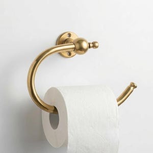 Könnte beinhalten: Ein goldfarbener, an der Wand befestigter Toilettenpapierhalter. Der Halter hat ein kreisförmiges Design mit einem gebogenen Arm zur Aufnahme der Rolle. Die Montageplatte ist rund mit Schraubenlöchern. Eine Rolle weißes Toilettenpapier ist eingesetzt.