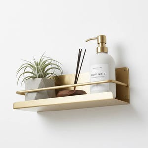 K&ouml;nnte beinhalten: Ein goldfarbenes Metallregal mit gebogener Frontschiene, das eine kleine Topfpflanze, eine Flasche Handseife und einen Holzdiffusor enth&auml;lt. Die Handseifenflasche hat eine goldene Pumpe und ein Etikett mit der Aufschrift "Scent No. 4 Citrus Bergamot".