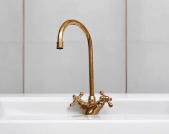 May include: A gold-colored faucet with a curved spout and cross-shaped handles. The faucet is mounted on a white sink, with a tiled wall in the background. The faucet is made of brass.