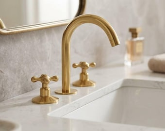 May include: A gold-toned bathroom faucet with a curved spout and two cross-shaped handles. The faucet is set against a marble countertop, with a matching mirror in the background. A bottle of perfume and a rolled towel are also visible.