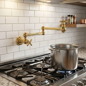 Könnte beinhalten: Ein goldfarbener, an der Wand montierter Topffüller-Wasserhahn mit kreuzförmigem Griff und Gelenkarm ist über einem Gasherd aus Edelstahl installiert. Ein Edelstahltopf steht auf dem Brenner, und weiße U-Bahn-Fliesen bedecken die Wand.