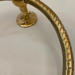 Close-up view of a gold-colored brass towel ring, The ring features a textured, rope-like design along its circular surface,  It's mounted on a circular base with visible screw holes, The ring's end is capped with a smooth, spherical detail, The overall style suggests a nautical or traditional design aesthetic, The brass has a polished, shiny finish