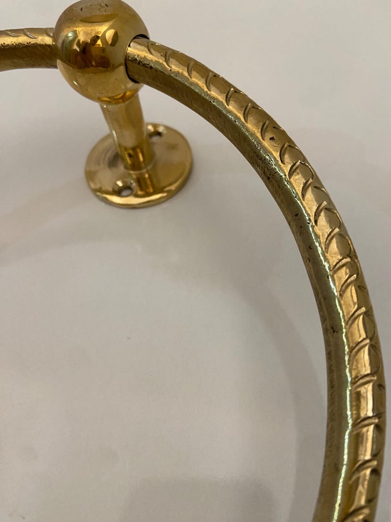 Close-up view of a gold-colored brass towel ring, The ring features a textured, rope-like design along its circular surface,  It's mounted on a circular base with visible screw holes, The ring's end is capped with a smooth, spherical detail, The overall style suggests a nautical or traditional design aesthetic, The brass has a polished, shiny finish