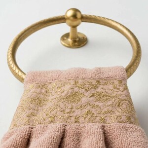 May include: A gold-colored towel ring holds a pink towel with a gold floral design. The towel ring is mounted on a white wall. The towel has a textured appearance.