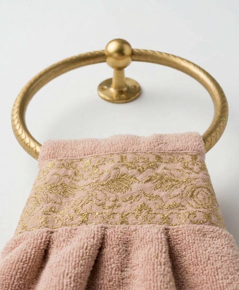 May include: A gold-colored towel ring holds a pink towel with a gold floral design. The towel ring is mounted on a white wall. The towel has a textured appearance.