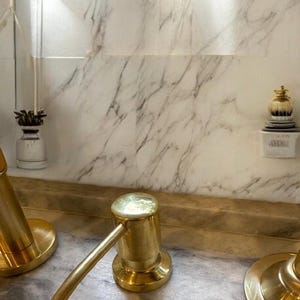 Könnte beinhalten: Nahaufnahme einer goldfarbenen Armatur mit Hebelgriff und rundem Sockel. Die Armatur befindet sich vor einem Hintergrund mit Marmormuster. Dekorative Elemente sind an der Wand sichtbar, darunter eine kleine Vase und ein dekoratives Objekt.