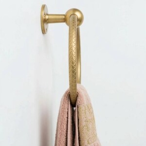 May include: A gold-colored towel ring with a textured design, mounted on a white wall. A pink towel with a gold embroidered border hangs from the ring. The towel ring has a circular design.