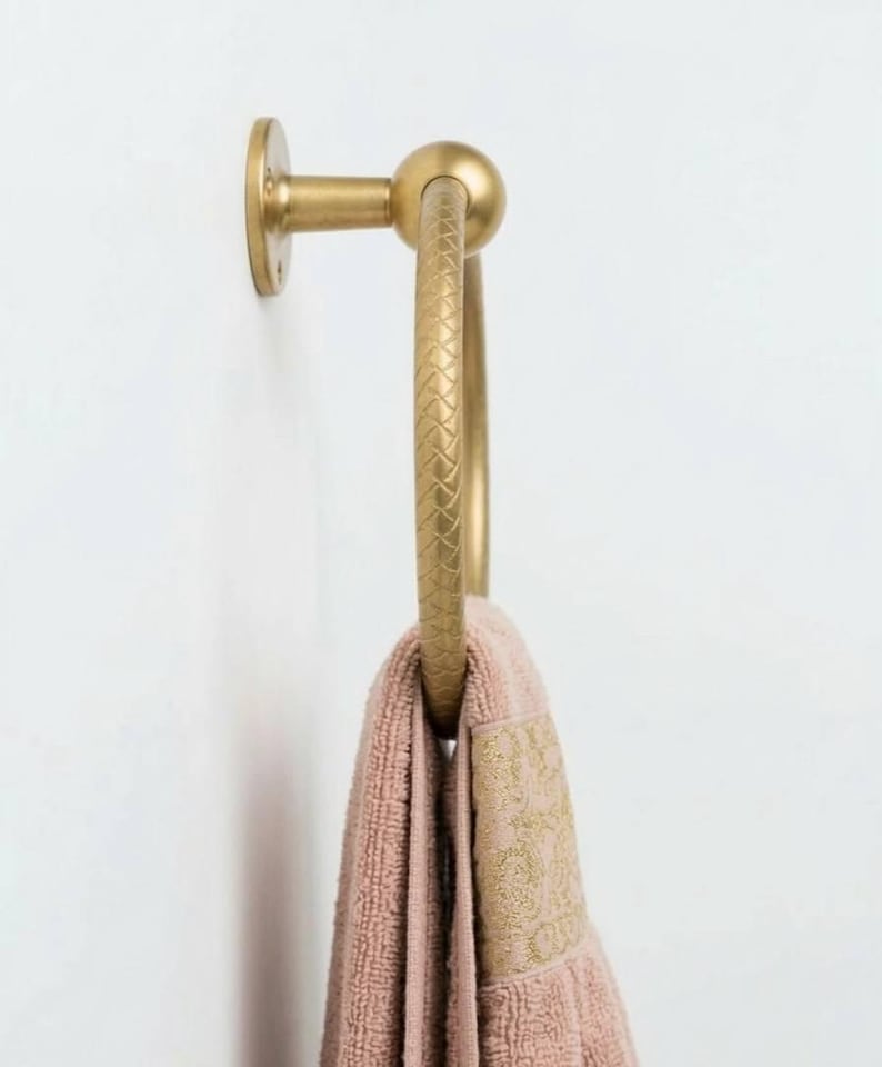 May include: A gold-colored towel ring with a textured design, mounted on a white wall. A pink towel with a gold embroidered border hangs from the ring. The towel ring has a circular design.