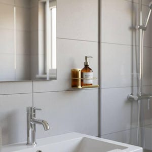 K&ouml;nnte beinhalten: Badezimmer mit wei&szlig;em Waschbecken und verchromtem Wasserhahn. Eine braune Glasflasche mit Handseife steht auf einem goldfarbenen Wandregal. Die W&auml;nde sind hellgrau gefliest. Spiegel und Dusche sind ebenfalls zu sehen.