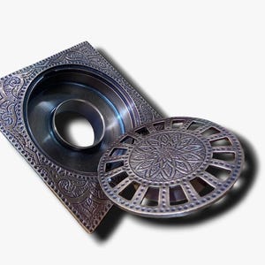 Könnte beinhalten: Ein quadratischer Abflussdeckel mit einem komplizierten Design und einem runden, dekorativen Gitter. Der Abflussdeckel ist aus Metall und hat eine Bronze-Oberfläche.