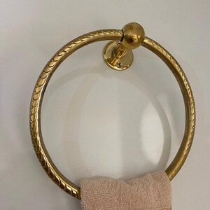 A gold-tone brass towel ring with a rope-like design hangs on a wall,  A light beige towel with gold embroidery is draped over the ring, The ring is circular with a small ball finial at the top, The wall-mounted design features a solid circular base