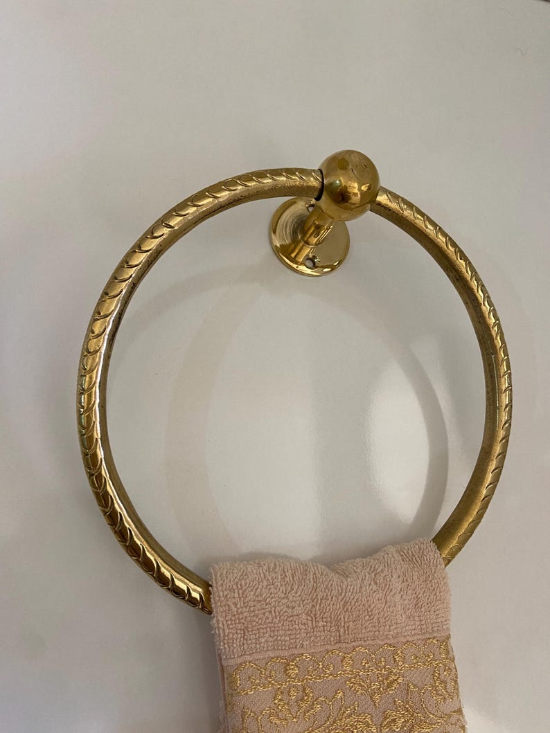 A gold-tone brass towel ring with a rope-like design hangs on a wall,  A light beige towel with gold embroidery is draped over the ring, The ring is circular with a small ball finial at the top, The wall-mounted design features a solid circular base