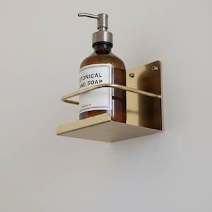 K&ouml;nnte beinhalten: Ein wandmontierter Seifenspenderhalter aus Messing mit einer braunen Glasflasche. Die Flasche hat eine silberne Pumpe und ein wei&szlig;es Etikett mit der Aufschrift "BOTANICAL AND SOAP". Der Halter hat ein kleines Regal und eine gebogene Stange.