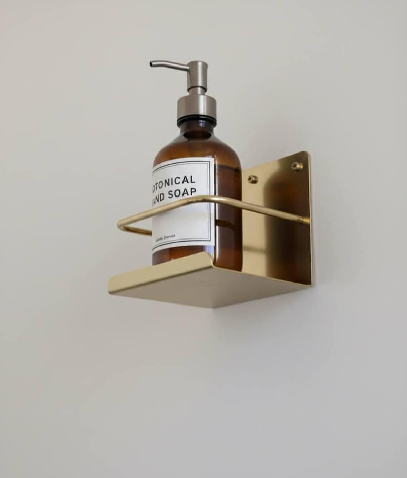 K&ouml;nnte beinhalten: Ein wandmontierter Seifenspenderhalter aus Messing mit einer braunen Glasflasche. Die Flasche hat eine silberne Pumpe und ein wei&szlig;es Etikett mit der Aufschrift "BOTANICAL AND SOAP". Der Halter hat ein kleines Regal und eine gebogene Stange.