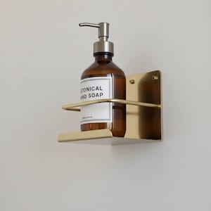 K&ouml;nnte beinhalten: Eine braune Glasflasche mit silbernem Pumpspender, beschriftet mit "BOTANICAL HAND SOAP", steht in einem goldfarbenen, an der Wand befestigten Metallhalter. Der Halter hat eine rechteckige R&uuml;ckplatte und eine gebogene Vorderseite. Das Design ist minimalistisch und modern.