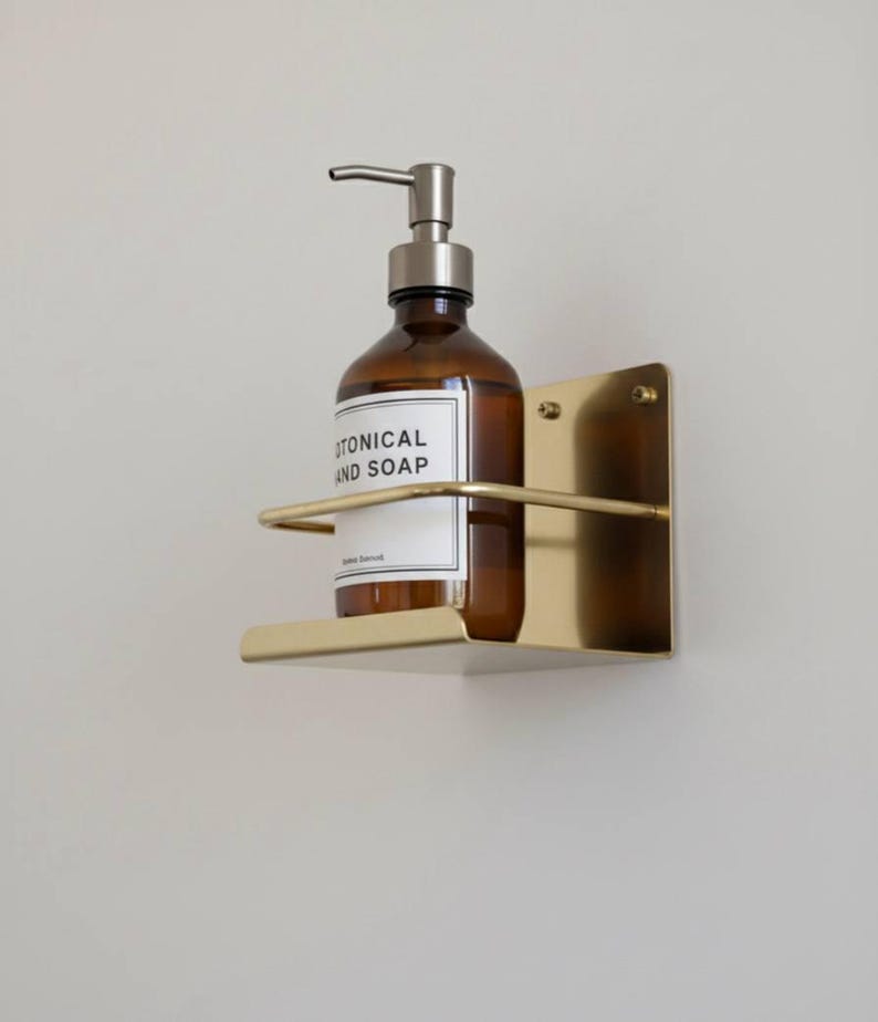 K&ouml;nnte beinhalten: Eine braune Glasflasche mit silbernem Pumpspender, beschriftet mit "BOTANICAL HAND SOAP", steht in einem goldfarbenen, an der Wand befestigten Metallhalter. Der Halter hat eine rechteckige R&uuml;ckplatte und eine gebogene Vorderseite. Das Design ist minimalistisch und modern.