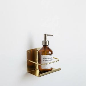 K&ouml;nnte beinhalten: Eine braune Glasflasche mit silbernem Pumpspender, beschriftet mit "BOTONICAL HAND SOAP", steht in einem goldfarbenen, an der Wand befestigten Metallhalter. Der Halter hat eine rechteckige Basis und eine gebogene Vorderseite. Der Hintergrund ist eine schlichte wei&szlig;e Wand.