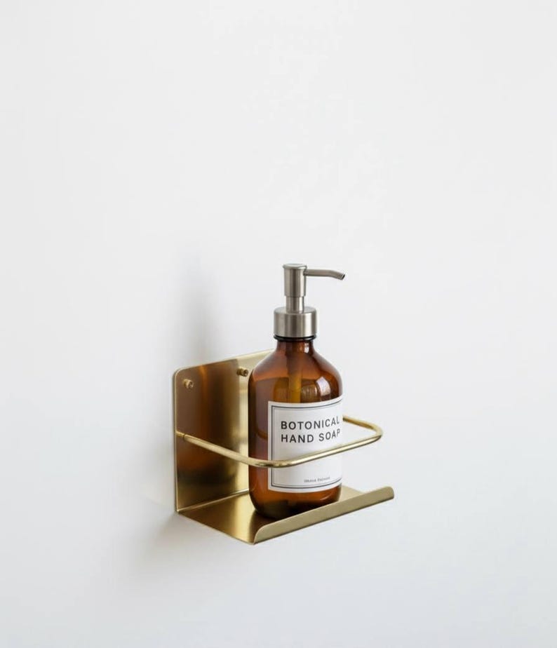 K&ouml;nnte beinhalten: Eine braune Glasflasche mit silbernem Pumpspender, beschriftet mit "BOTONICAL HAND SOAP", steht in einem goldfarbenen, an der Wand befestigten Metallhalter. Der Halter hat eine rechteckige Basis und eine gebogene Vorderseite. Der Hintergrund ist eine schlichte wei&szlig;e Wand.